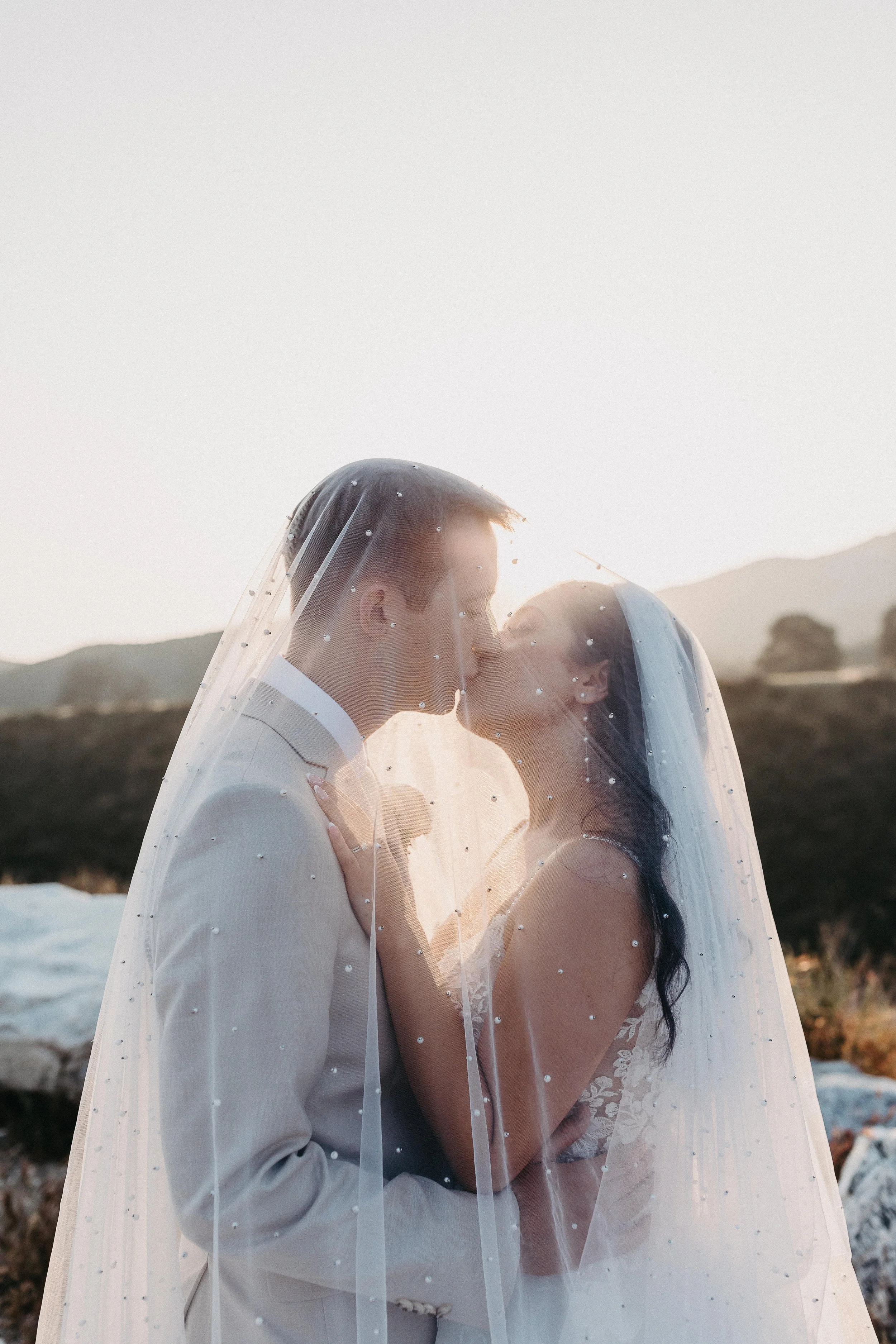 Bride and groom kissing under a pearl beaded veil during golden hour.