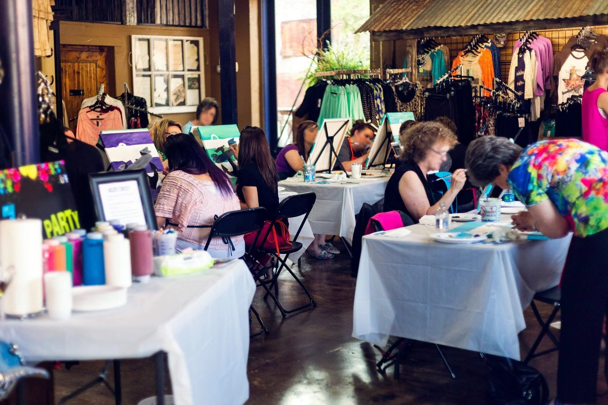 Women participating in a painting class inside a boutique with clothing displays in the background.