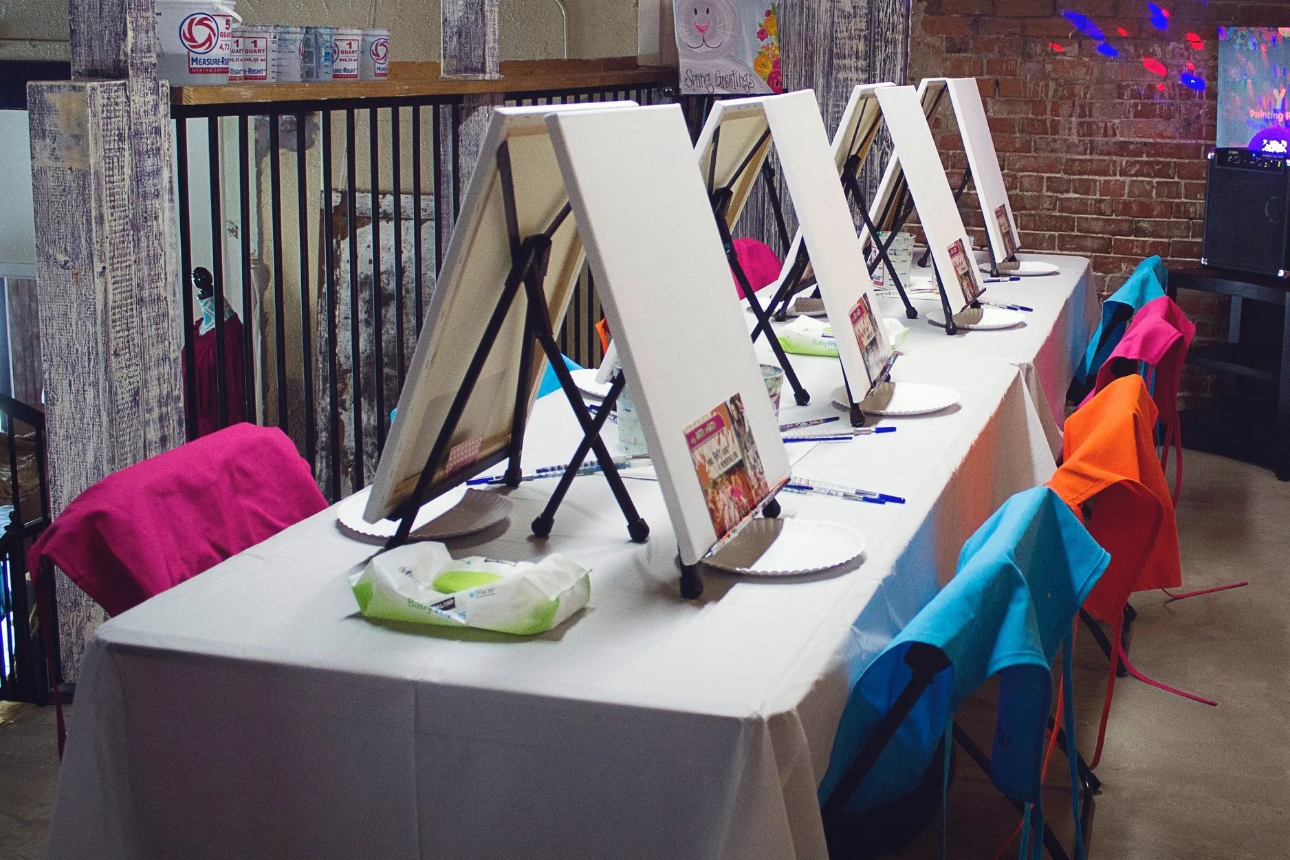 A table set up for a painting activity with multiple blank canvases on small easels, each accompanied by a paper plate, a marker, and a promotional card, with a seating of colorful chairs draped in cloth, against a background featuring a brick wall and a sunny greetings poster.