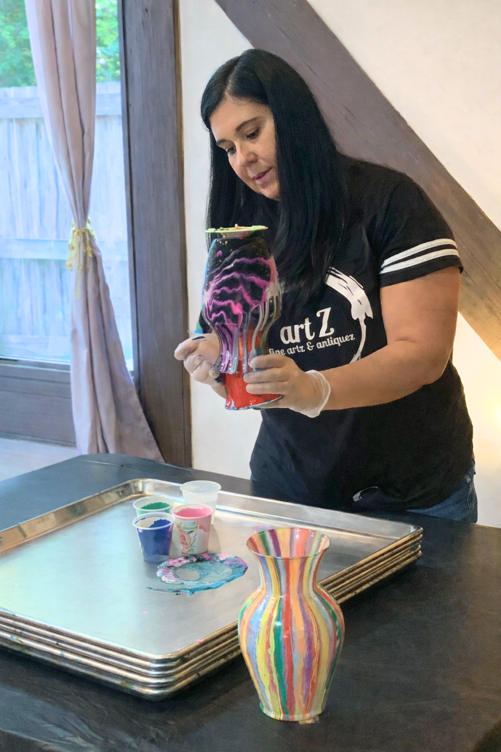 Woman holding a colorful art glass vase in a room with a window, wooden paneling, and a table with small paint containers and a rainbow-colored ceramic vase.