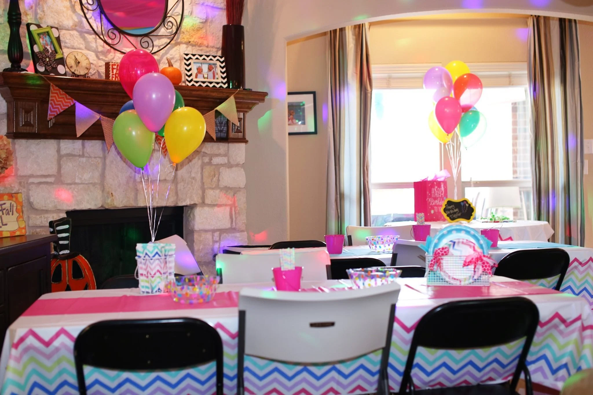 Colorful birthday party setup with tables decorated with chevron tablecloths, pink cups, and bowls, and balloons in pink, purple, yellow, and green tied to centerpieces. Balloons are also grouped in the background near a window with natural light.