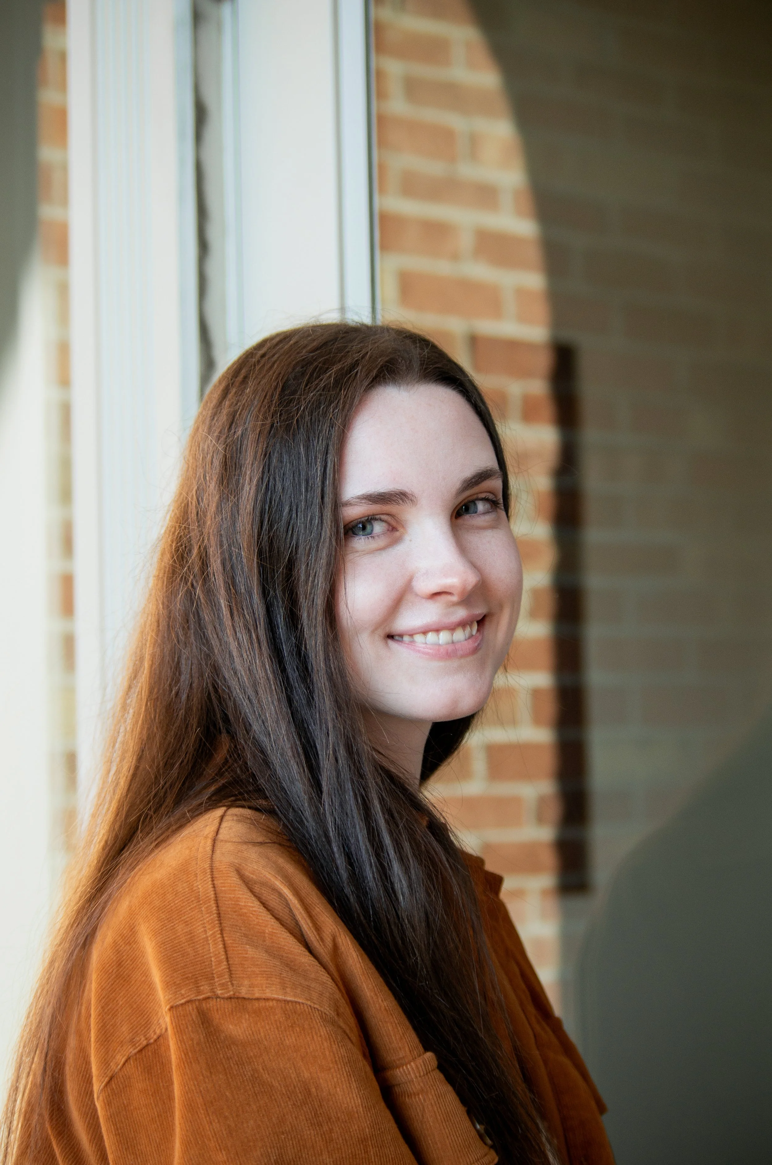 A young woman with long brown hair and blue eyes smiling, standing near a window with a brick wall in the background.