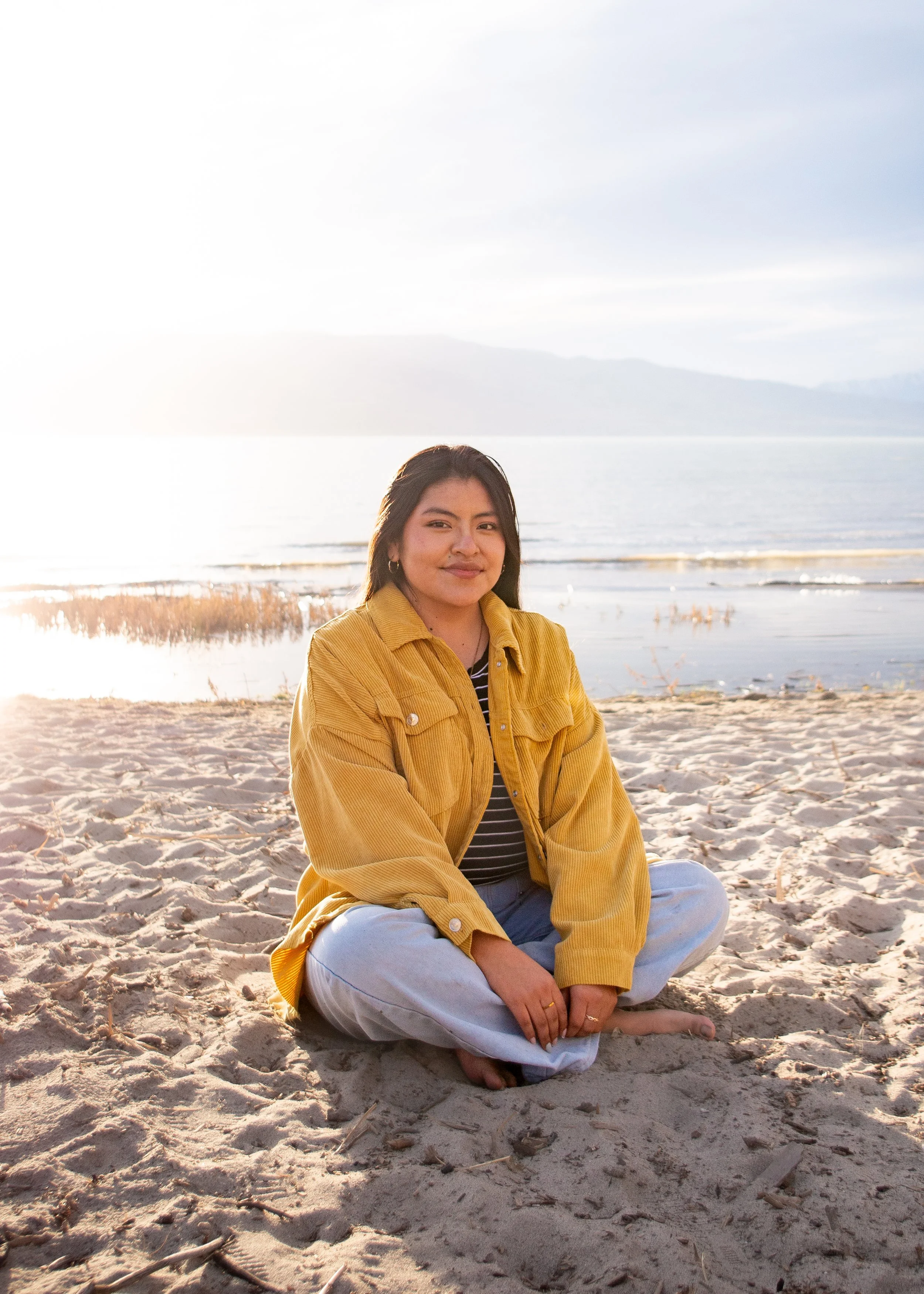 A woman with dark hair sits cross-legged on the sandy beach near a body of water, wearing a yellow jacket and smiling at the camera during sunset