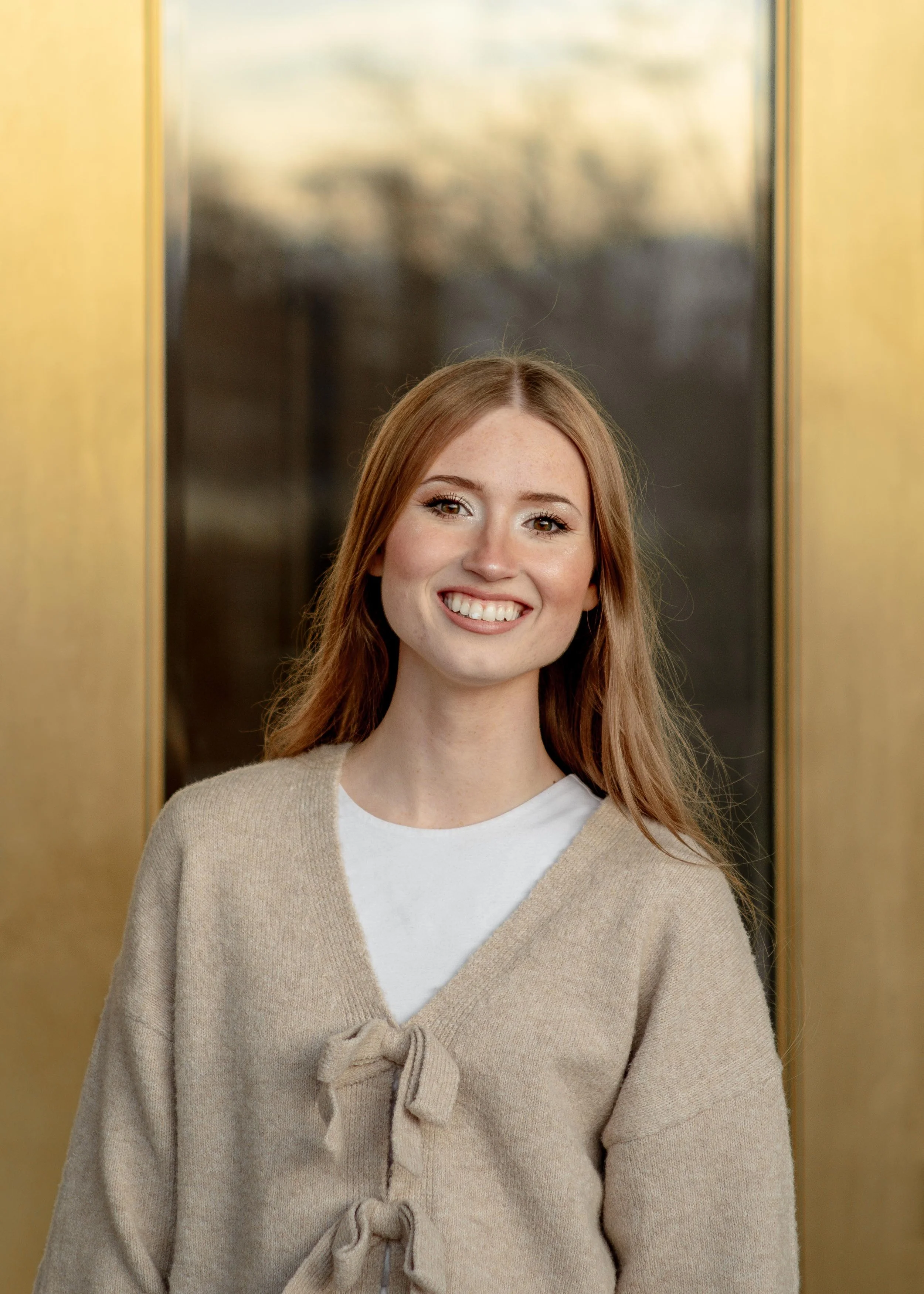 Young woman with red hair smiling, wearing a beige cardigan with bows over a white shirt, standing in front of an elevator door.