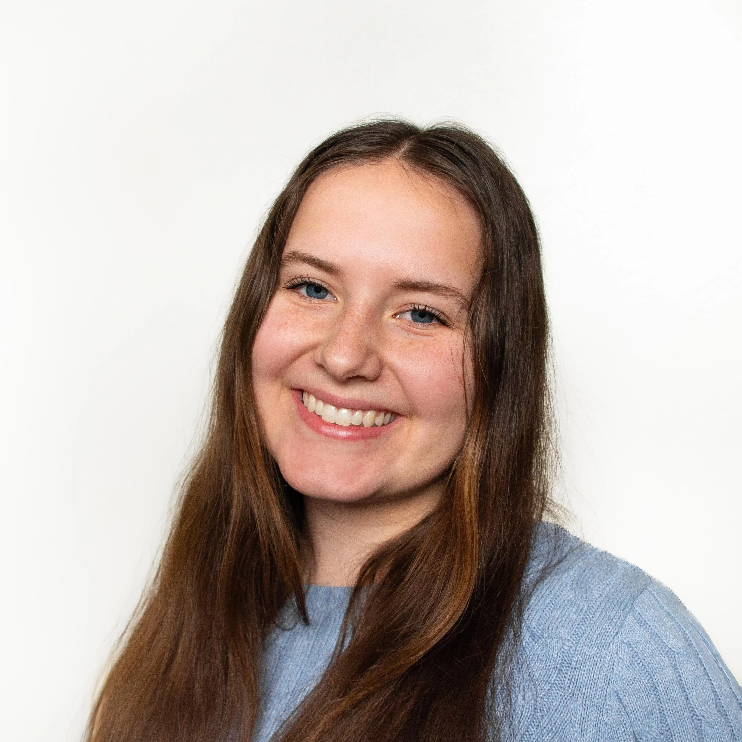 Close-up of a smiling woman with long brown hair, wearing a light blue sweater, against a plain white background.