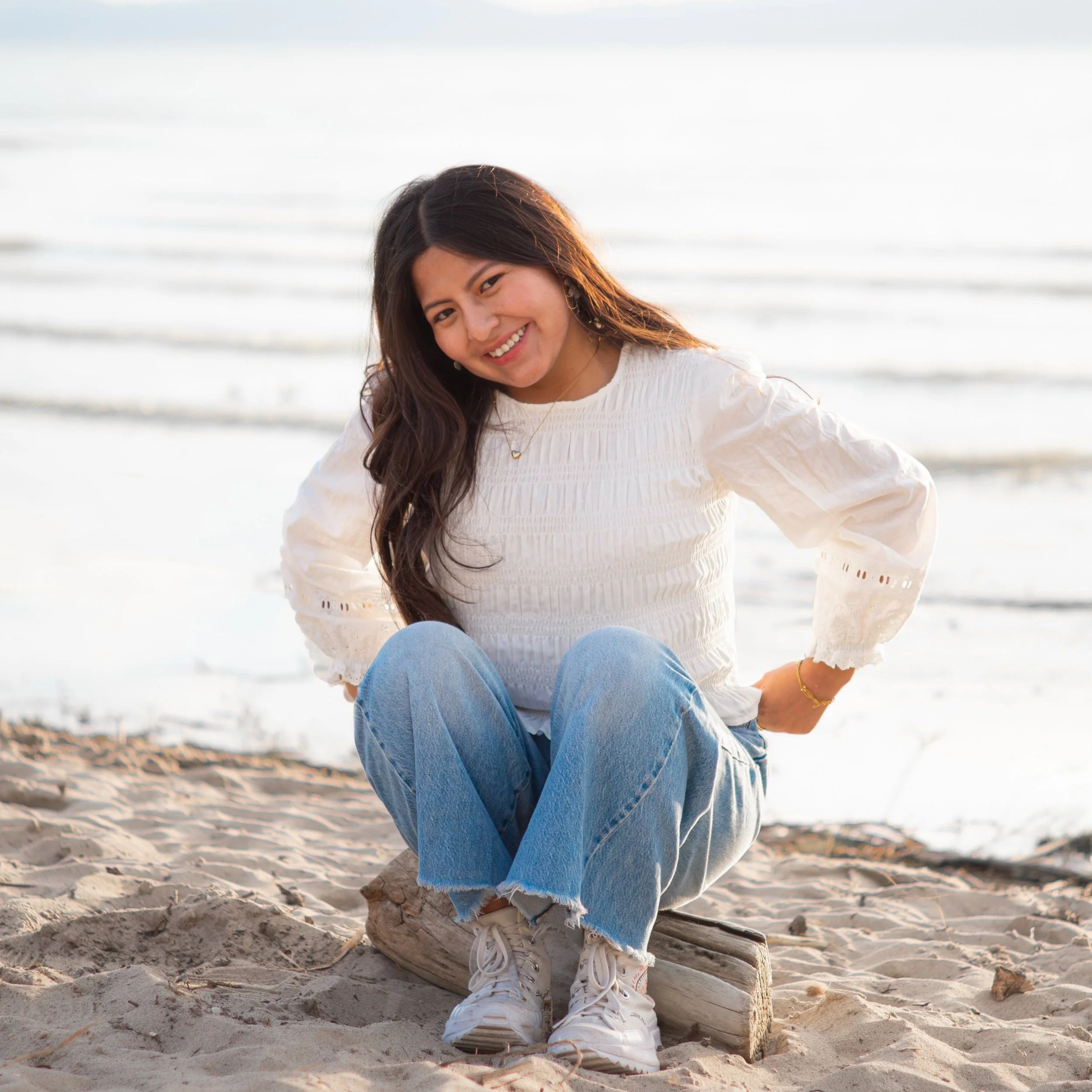 Young woman sitting on a log at the beach, smiling at the camera, with the ocean in the background.