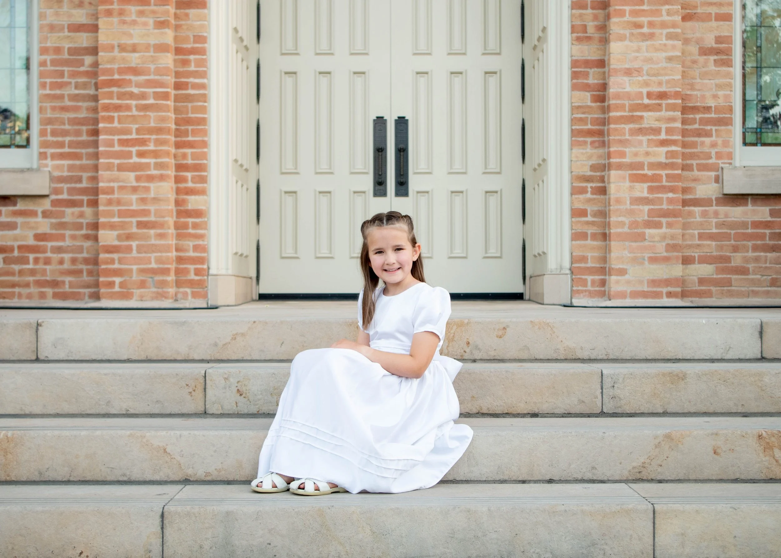 A young girl in a white dress sitting on the front steps of a brick house with a white door.