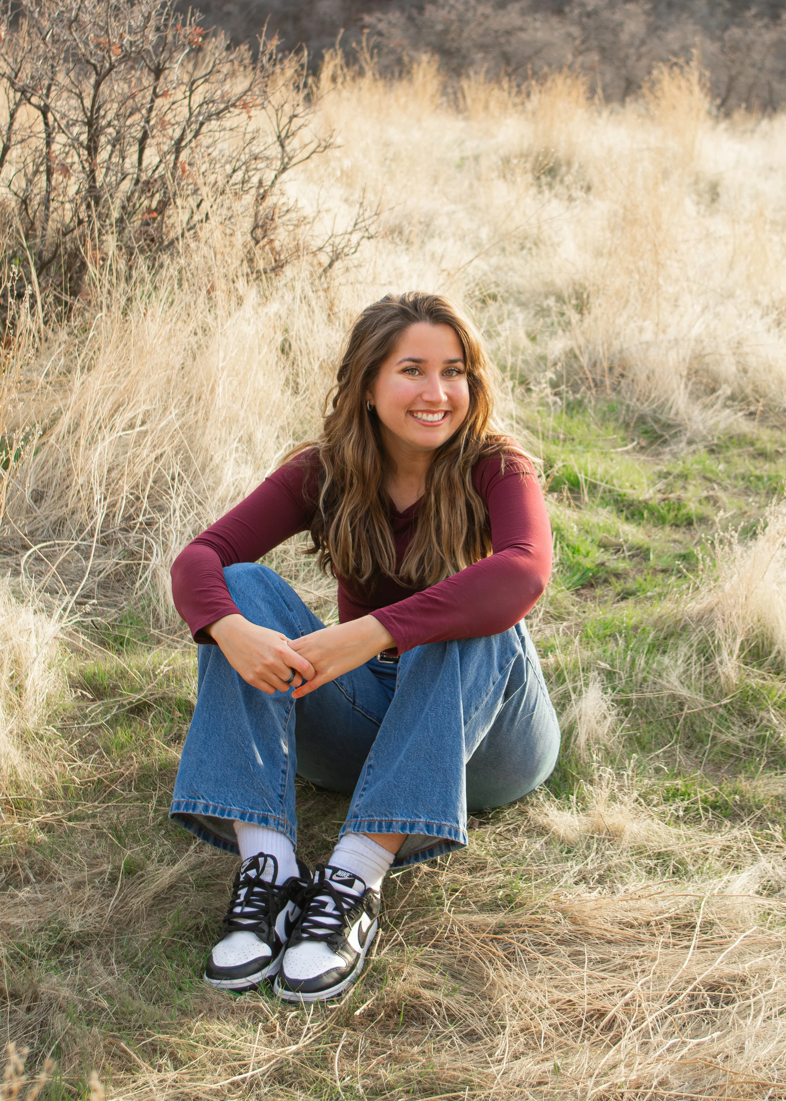 A young woman with long brown hair smiling and sitting on the ground in a grassy outdoor field, wearing a maroon long sleeve shirt, blue jeans, and football sneakers. The background features dried grass, green plants, and some bushes.