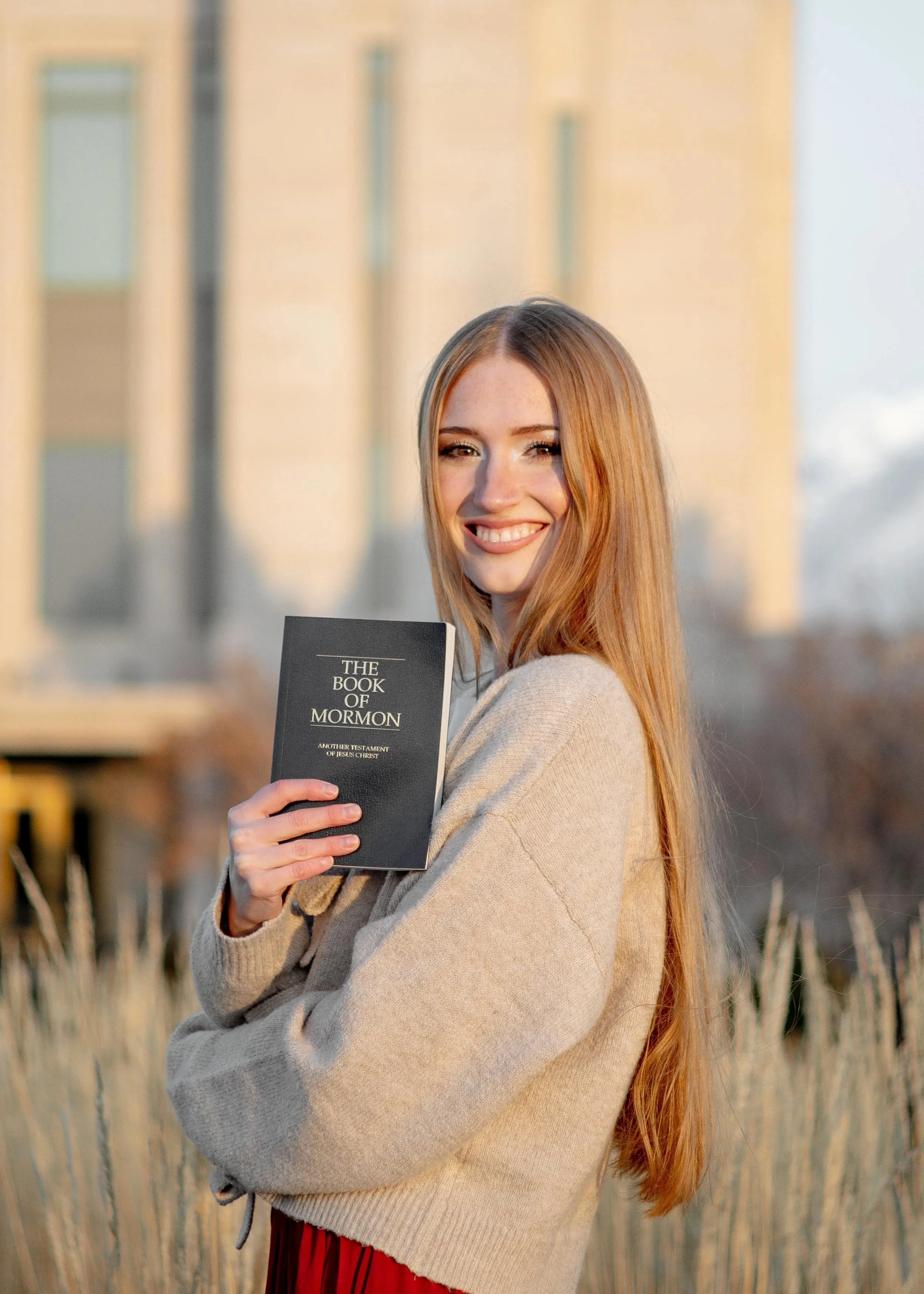 A young woman with long red hair smiling and holding a black book titled 'The Book of Mormon' outdoors in front of a building with tall grasses in the foreground.