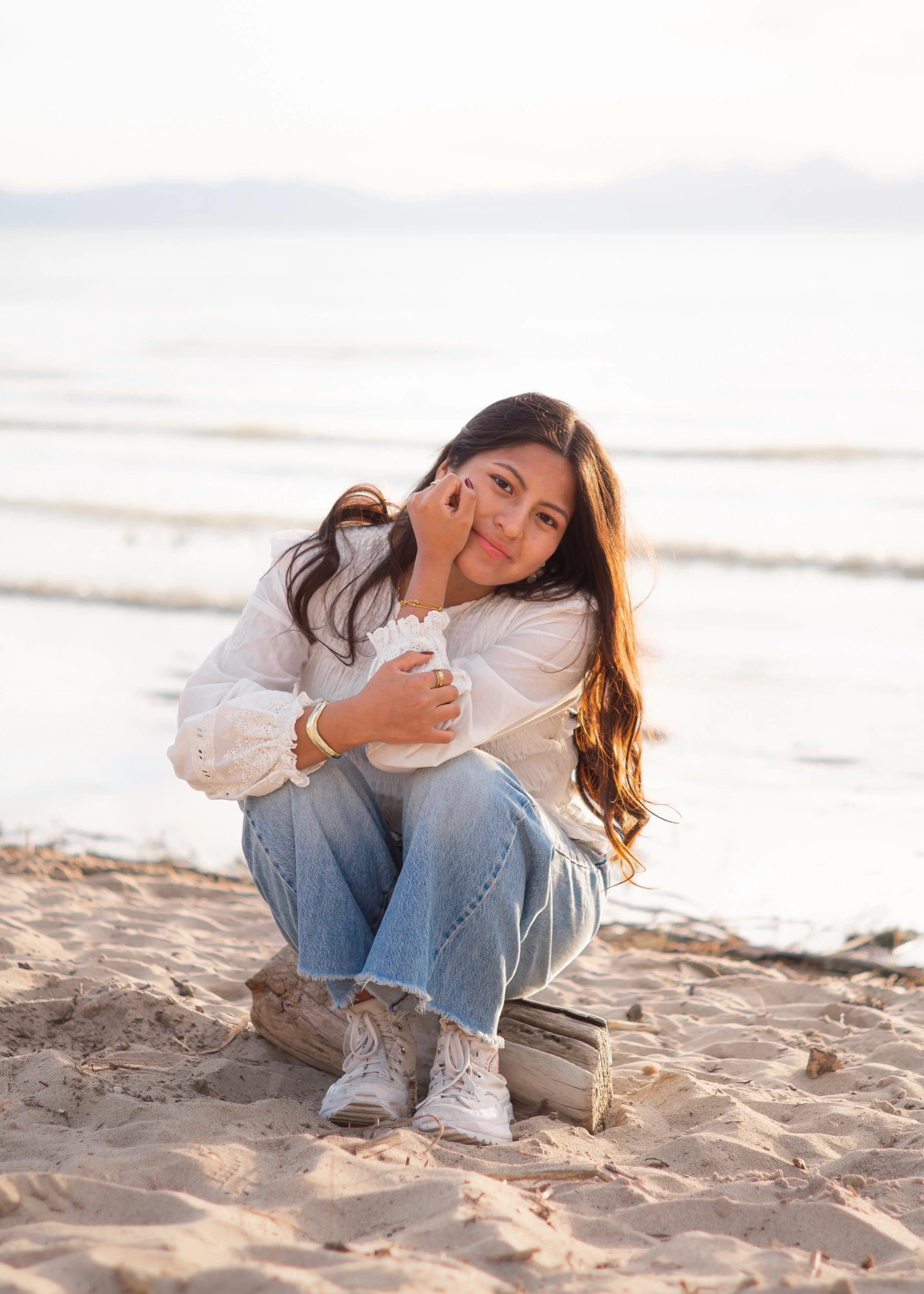 Young woman sitting on a log at the beach, smiling at the camera, with the ocean in the background.