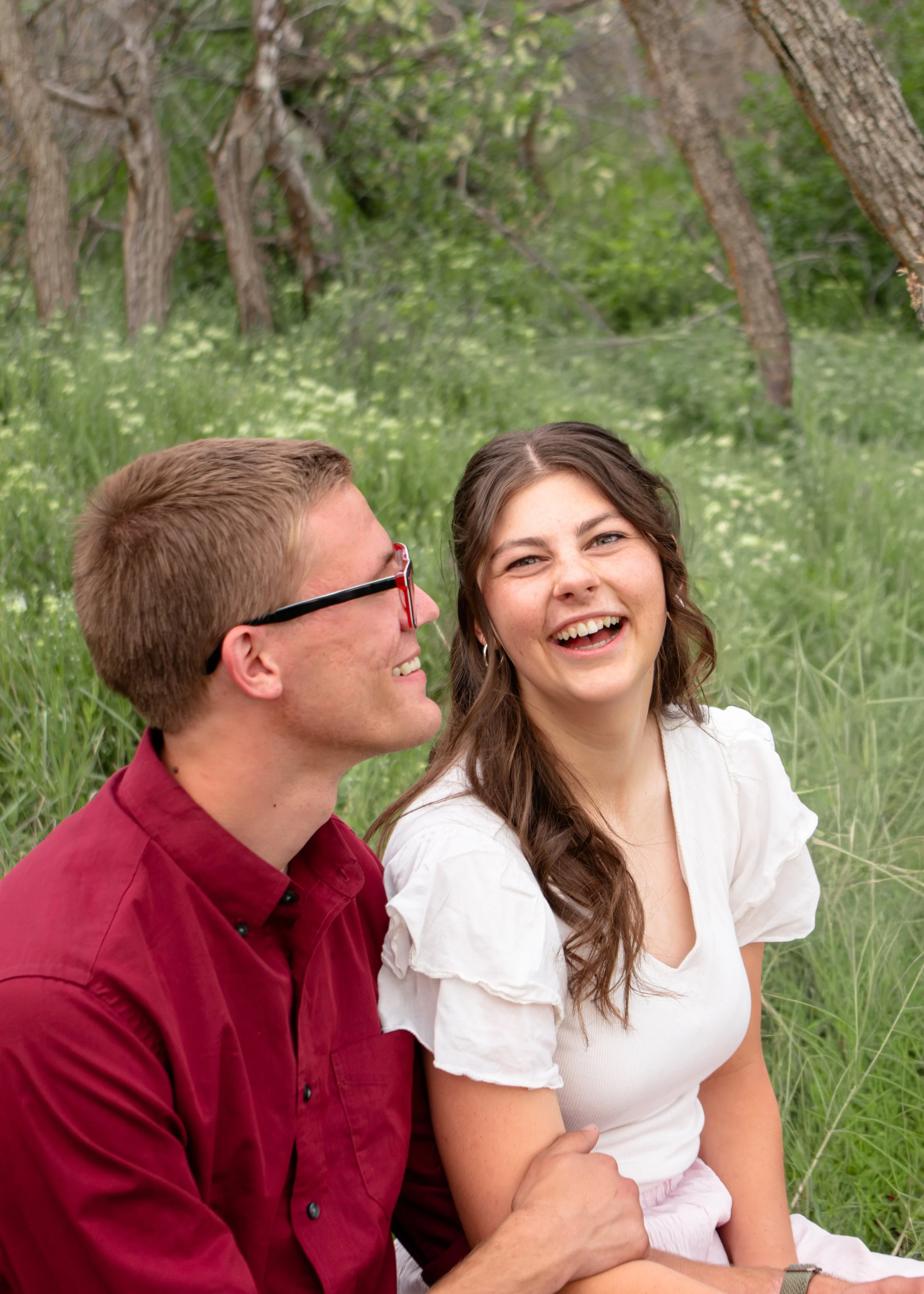 A man with glasses and a woman sitting outdoors in a green, wooded area, smiling and looking at each other.