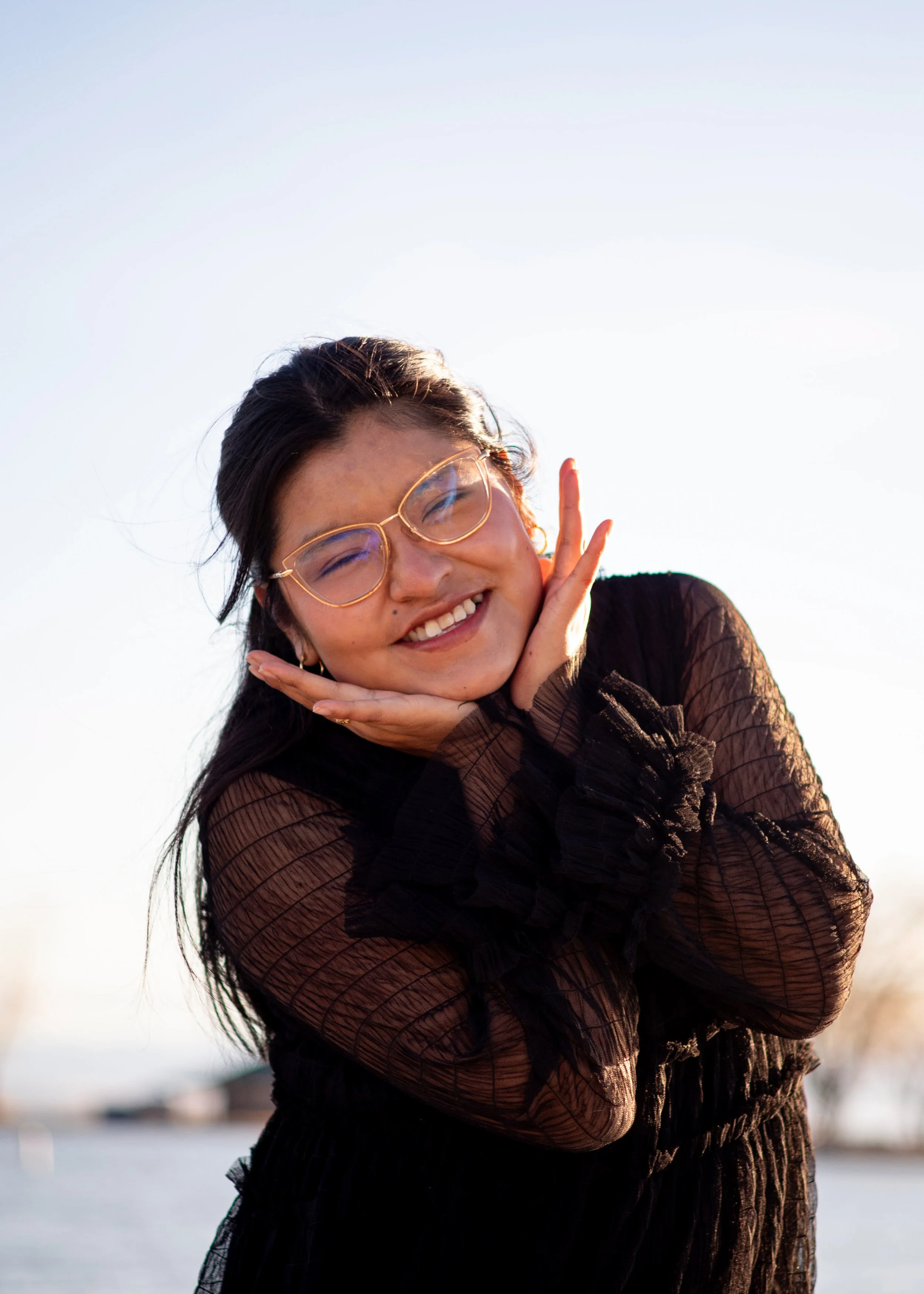 A young woman smiling with glasses, posing with her hands on her face outdoors during daytime.
