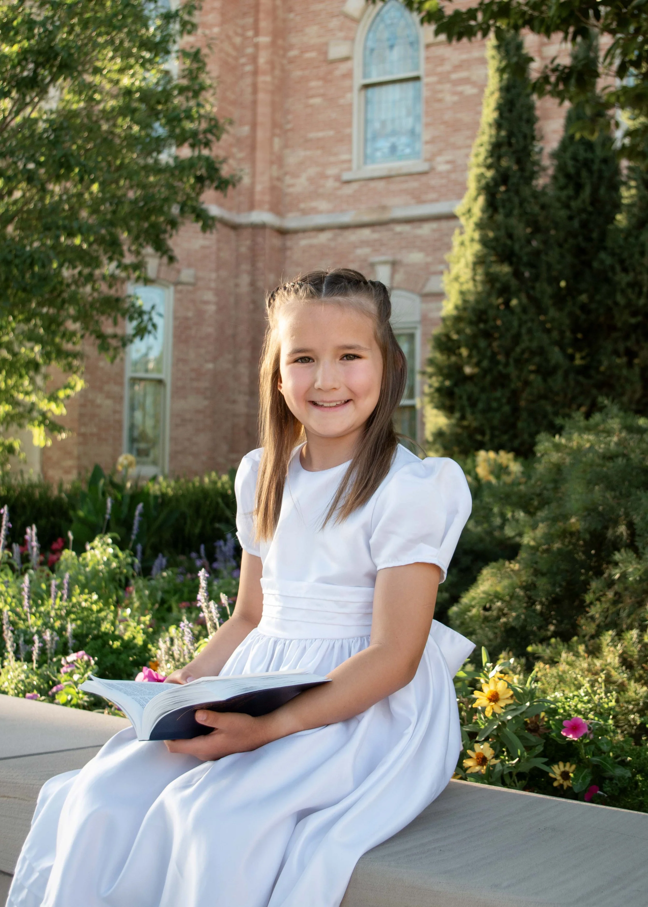 A young girl in a white dress sitting outdoors on a bench, holding an open book, smiling, with a brick church and greenery in the background.