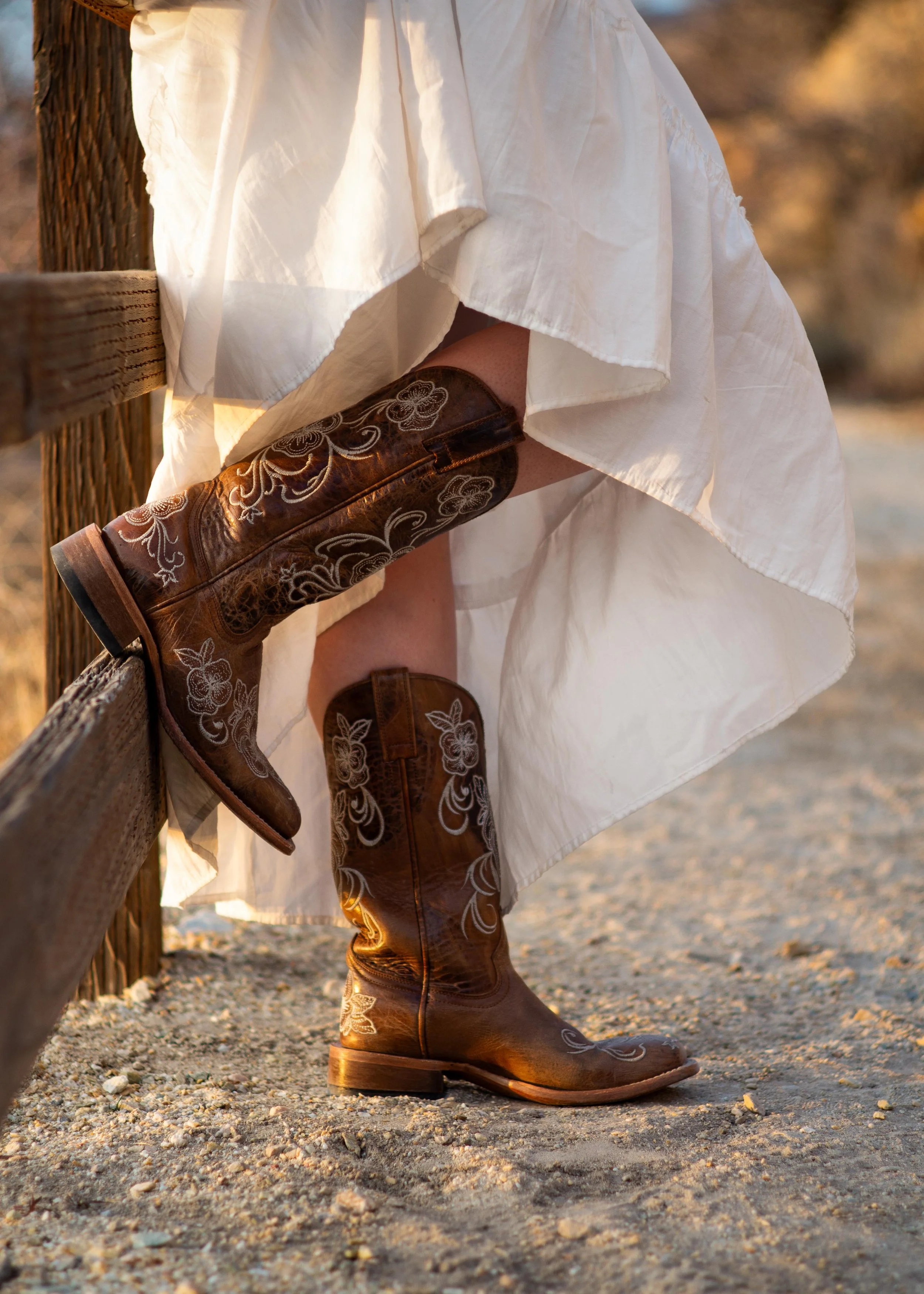 A person wearing brown embroidered cowboy boots, with a white flowing dress, sitting on a wooden fence in a desert landscape at sunset.