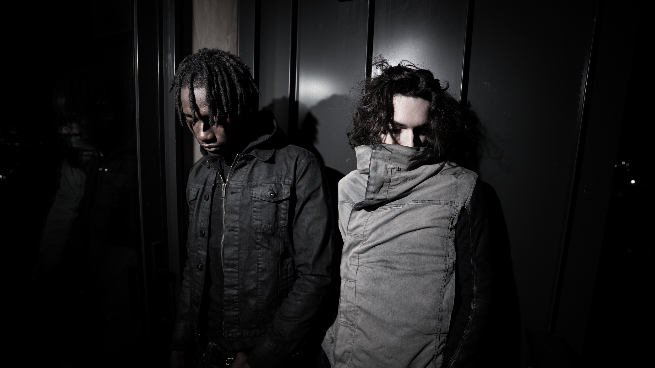 A young man with dreadlocks and a young woman with curly hair stand next to each other against a dark background, looking down with serious expressions.