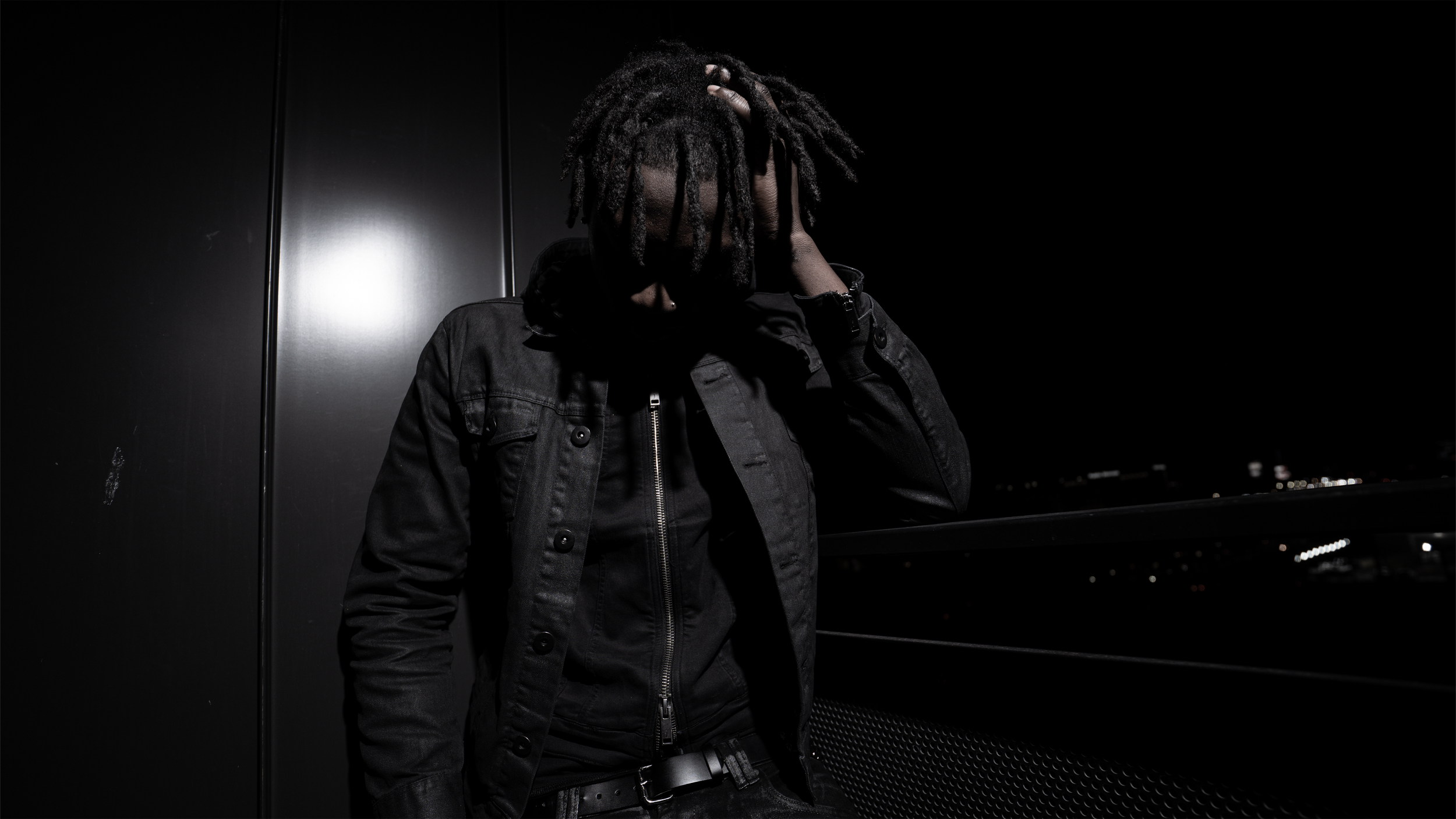 A person with dreadlocks dressed in dark clothing, standing on a balcony at night, with his hand on his head, in a dimly lit environment.