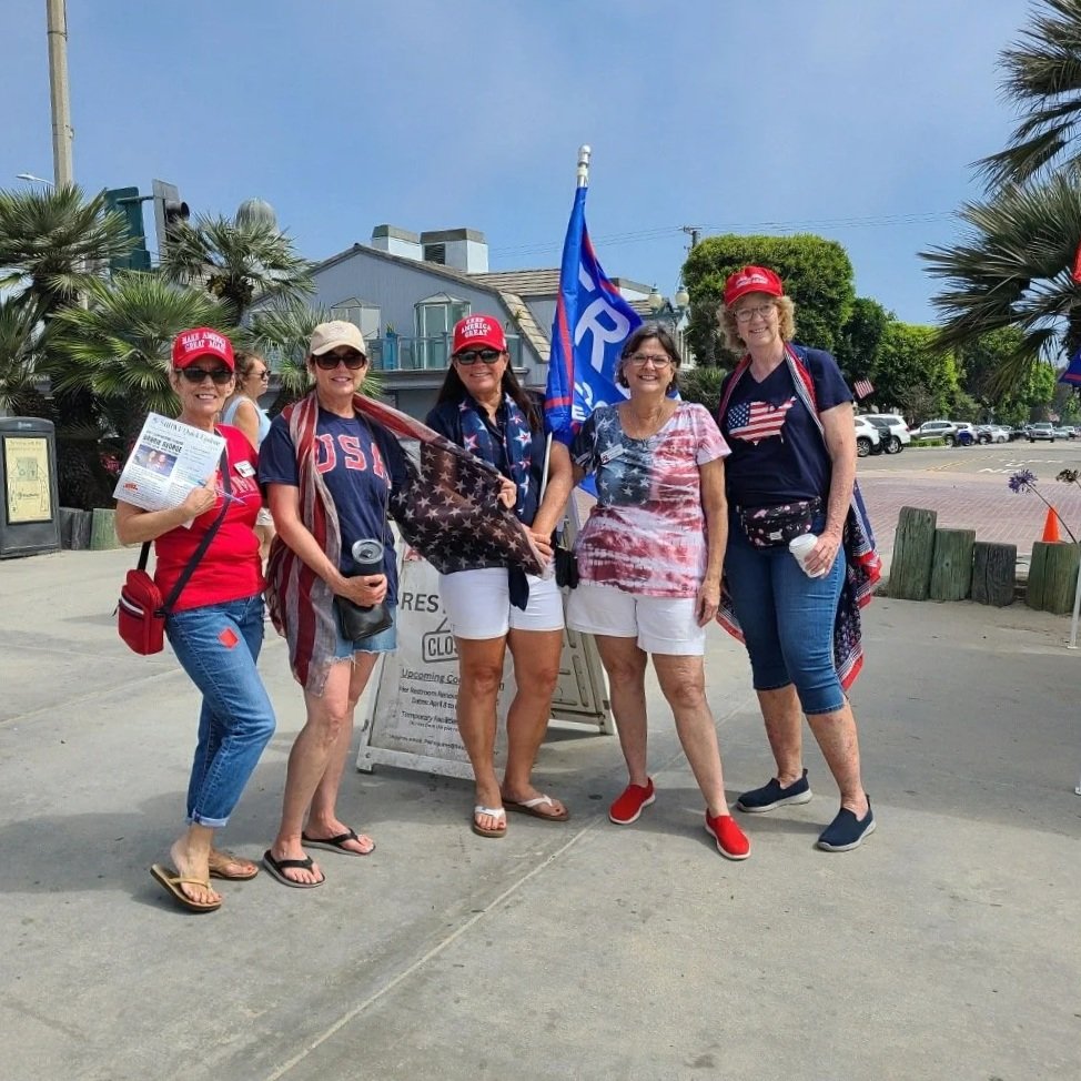 Five women standing together outdoors, dressed in patriotic red, white, and blue clothing, holding American flags and Trump flags, smiling for the photo. Some are wearing hats supporting Donald Trump.