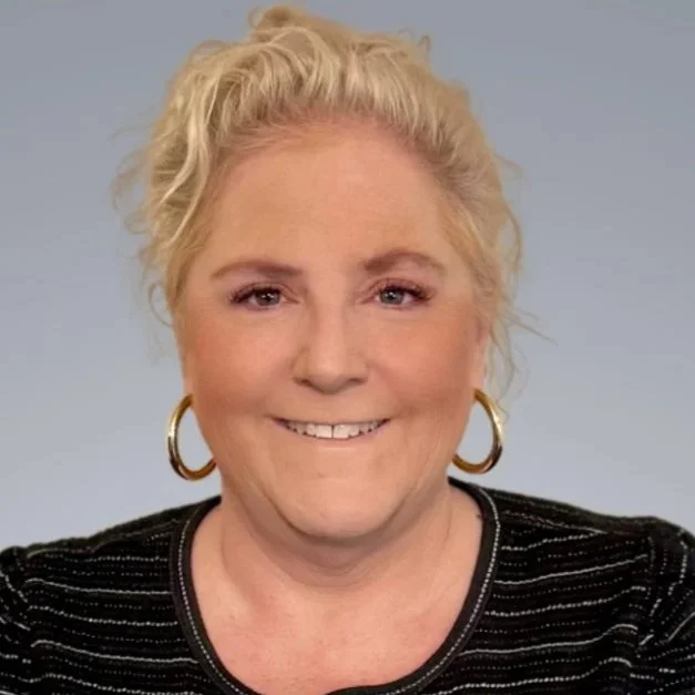 Close-up portrait of a smiling woman with blonde hair in a loose bun, wearing gold hoop earrings and a black top with silver thread details, against a plain light gray background.