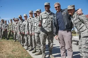 Group of military personnel in uniform standing in a line outdoors, with two men in civilian clothing in front, posing for a photo.