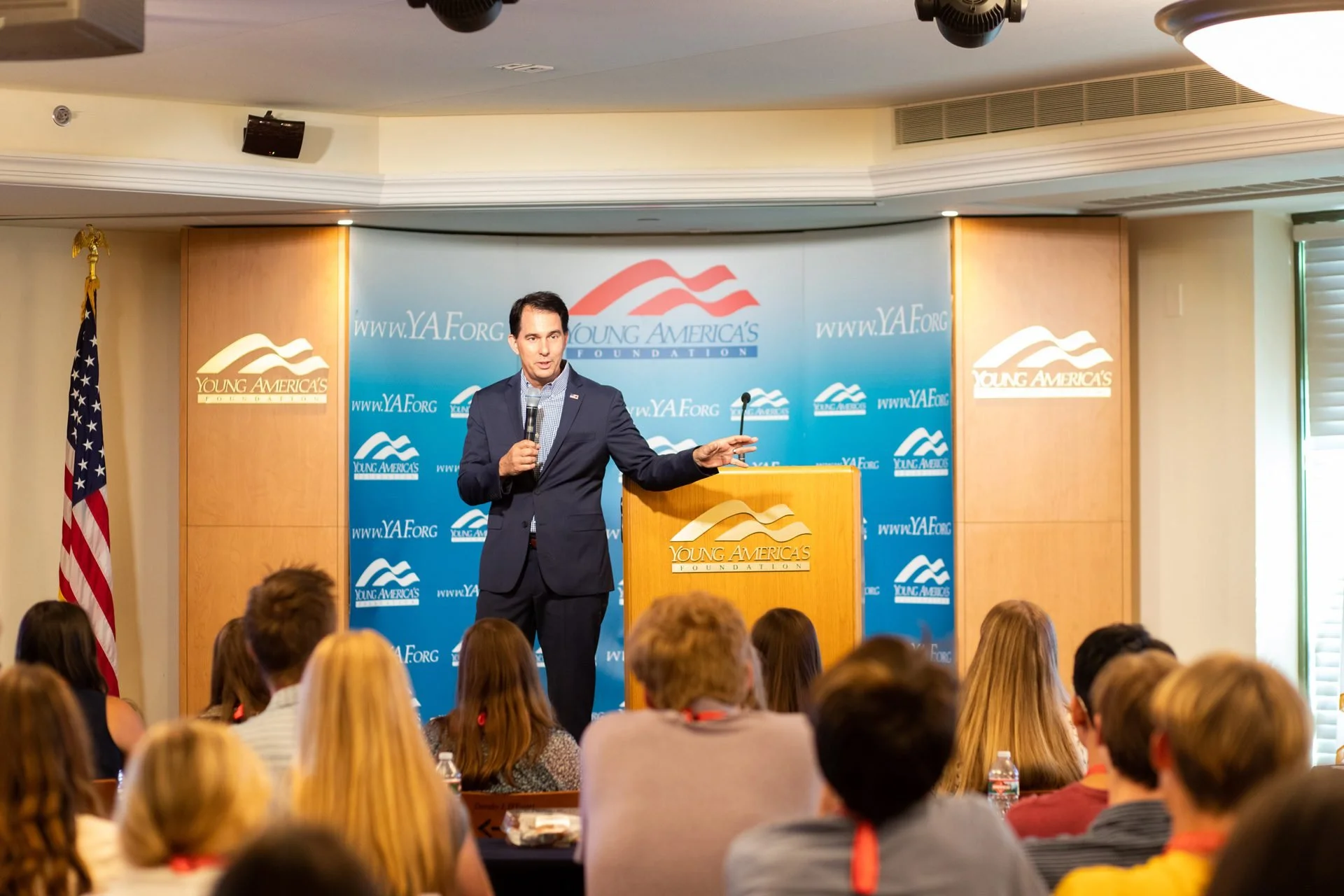 A man in a suit standing at a podium with a microphone, addressing an audience at a Young America's Foundation event with a blue backdrop and American flag to the side.