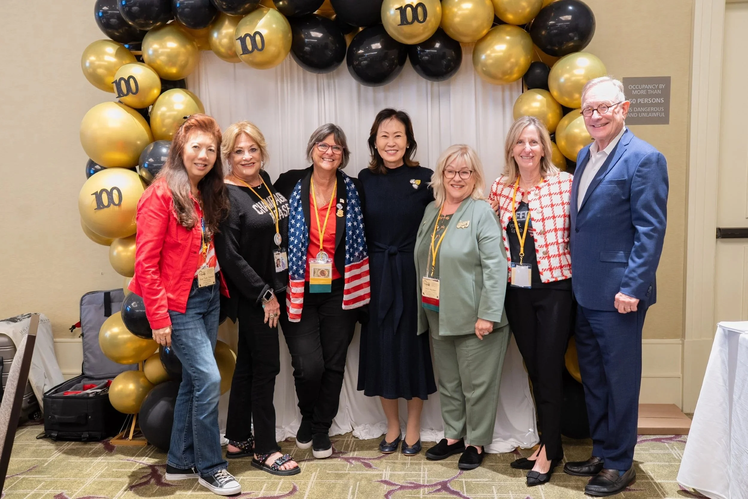 Group of eight diverse people standing together in front of a photo backdrop decorated with black and gold balloons, celebrating a 100th anniversary event.