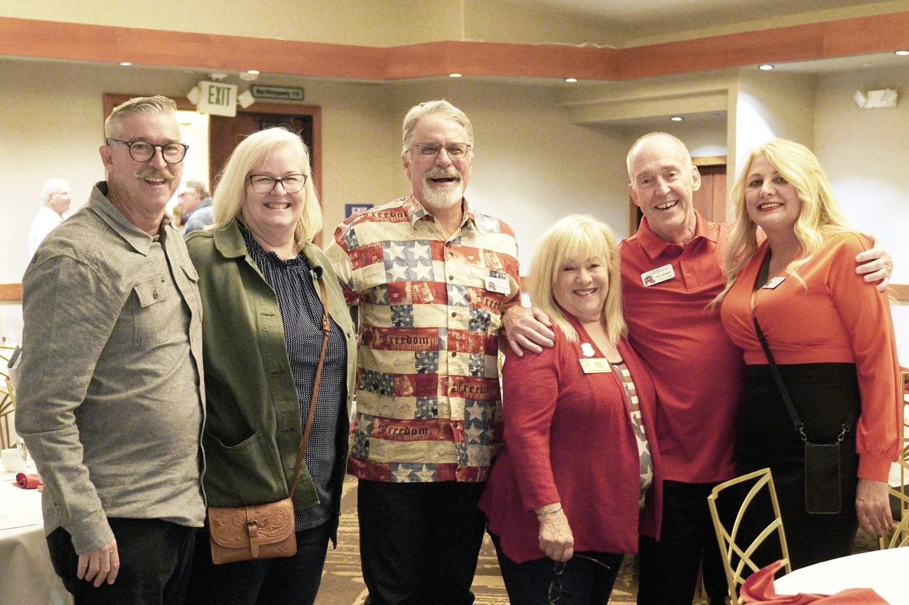 Group of six smiling adults standing together in a banquet hall, with four men and two women, some wearing glasses, and one woman wearing a red blazer, all posing for a photo.