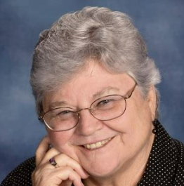 An elderly woman with gray hair, glasses, and a warm smile, wearing a black polka dot top and a ring, set against a blue background.