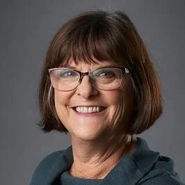 Smiling middle-aged woman with shoulder-length brown hair, glasses, and a teal top, posing against a gray background.