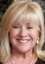 Close-up of a smiling woman with blonde hair, wearing pearl earrings and a pearl necklace.