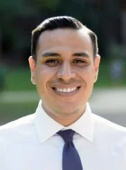 A smiling man with dark hair, wearing a white shirt and a dark tie, outdoors in a park-like setting.