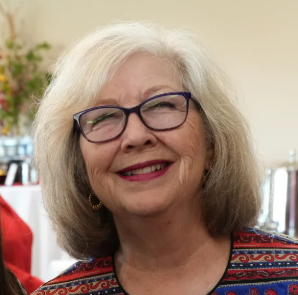 A smiling elderly woman with gray hair and glasses, wearing a colorful patterned top, in an indoor setting.