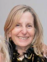 A smiling woman with blonde hair, wearing a black outfit with gold accents.