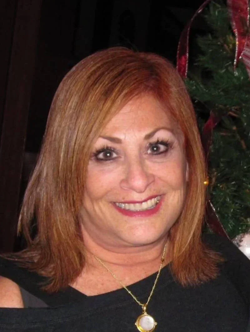 A woman with shoulder-length red hair, wearing makeup, a black top, a gold necklace with a large pendant, and smiling, standing near a Christmas tree with red ribbon decorations.