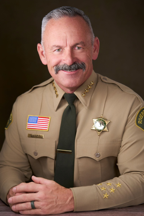 A smiling man in a sheriff uniform with a badge, American flag patch, and four gold stars on the shoulder, sitting with hands folded.