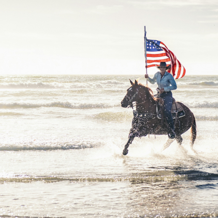 A person riding a horse through shallow water on a beach, holding an American flag.