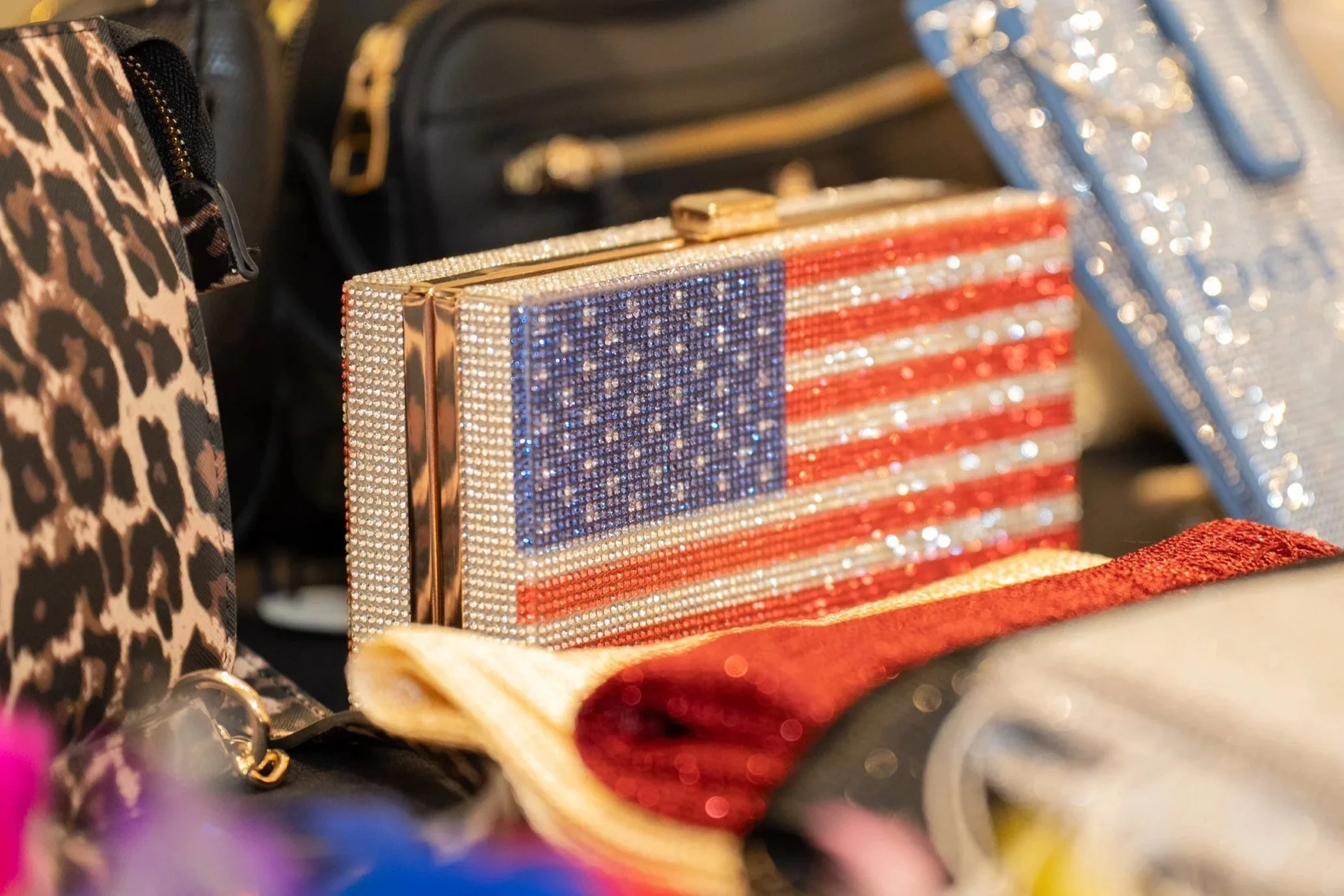A handbag decorated with rhinestones displaying the American flag, surrounded by other bags and accessories.