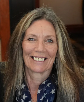 A woman with long blonde hair, smiling, wearing a blue top with white stars, in an indoor setting.
