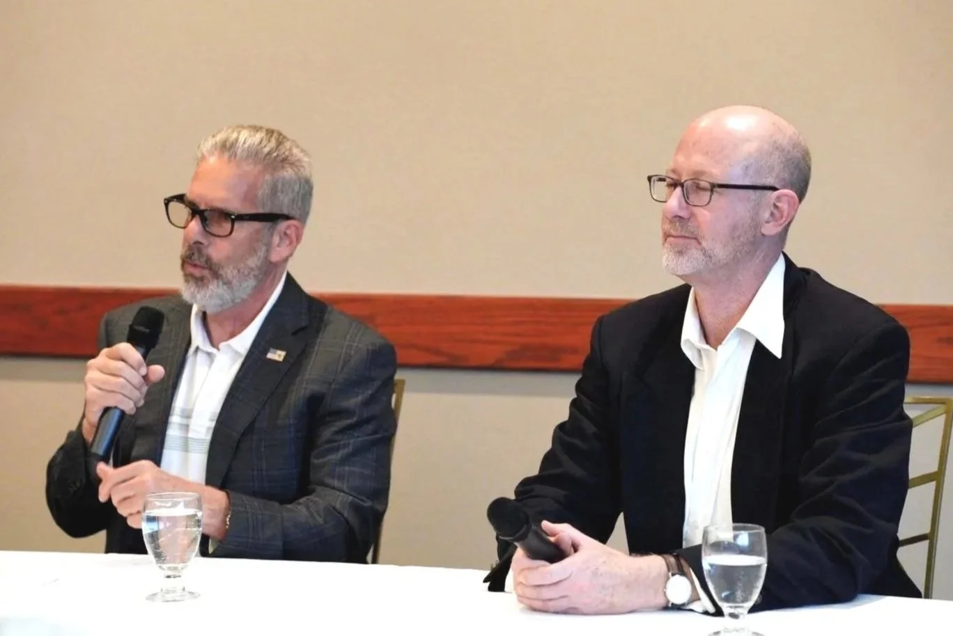 Two middle-aged men with glasses and gray beards sitting at a table during a panel or press conference. One is speaking into a microphone, and both have glasses of water in front of them.