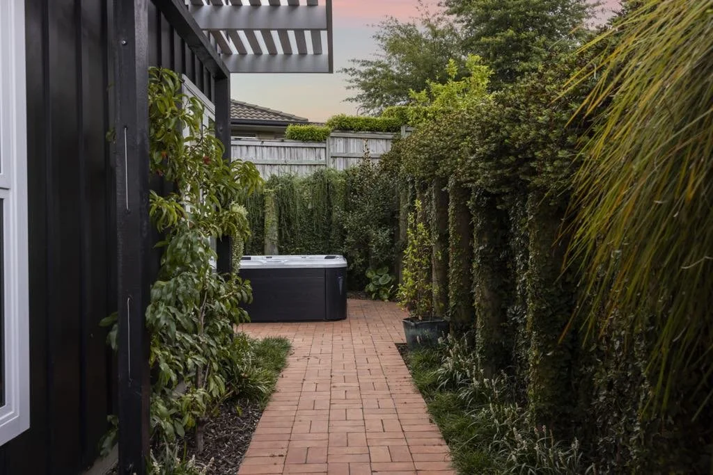 A narrow brick pathway in a backyard, lined with green bushes and plants on both sides, with a hot tub at the end of the path, and a wooden fence at the back under a pink and purple sky.