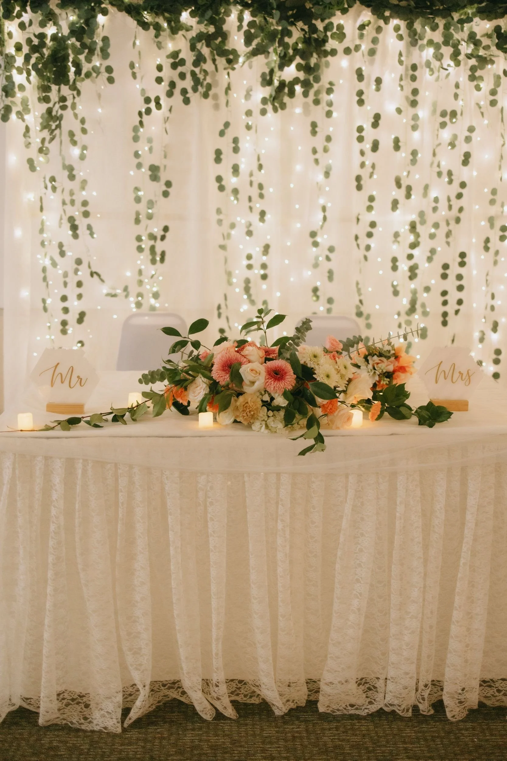Wedding reception table with a floral centerpiece, candles, and signs reading 'Mr' and 'Mrs', set against a backdrop of hanging greenery and string lights.