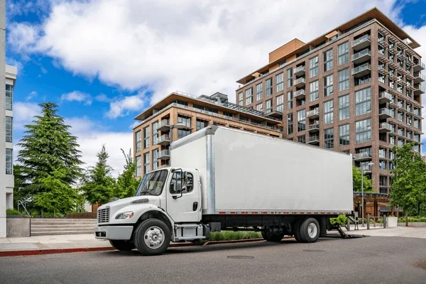 White delivery truck parked on the street in front of modern apartment buildings with trees and a partly cloudy sky.
