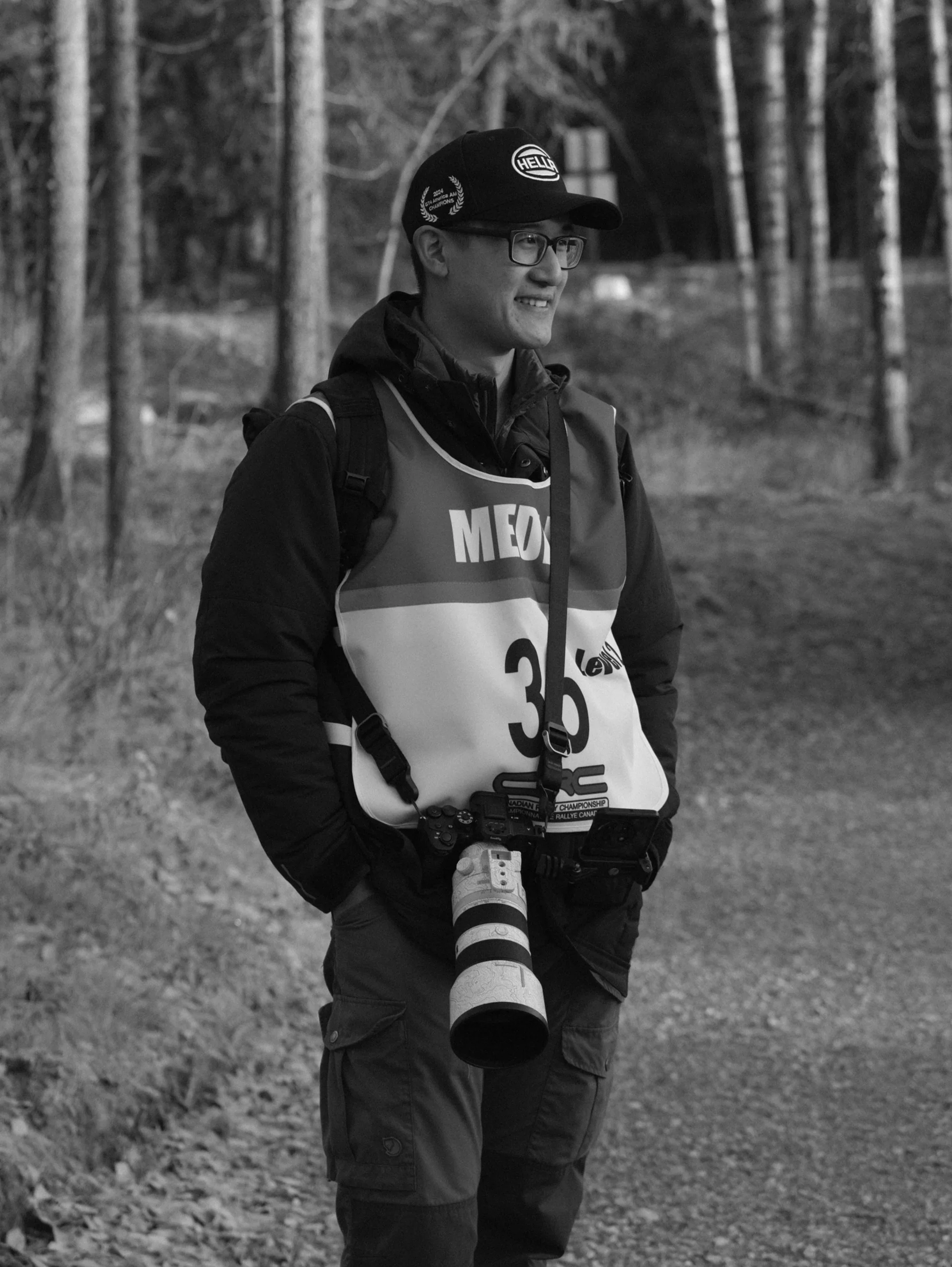 A man wearing glasses, a baseball cap, a vest with the number 36 on it, and a camera with a large telephoto lens around his neck, standing outdoors with trees in the background.