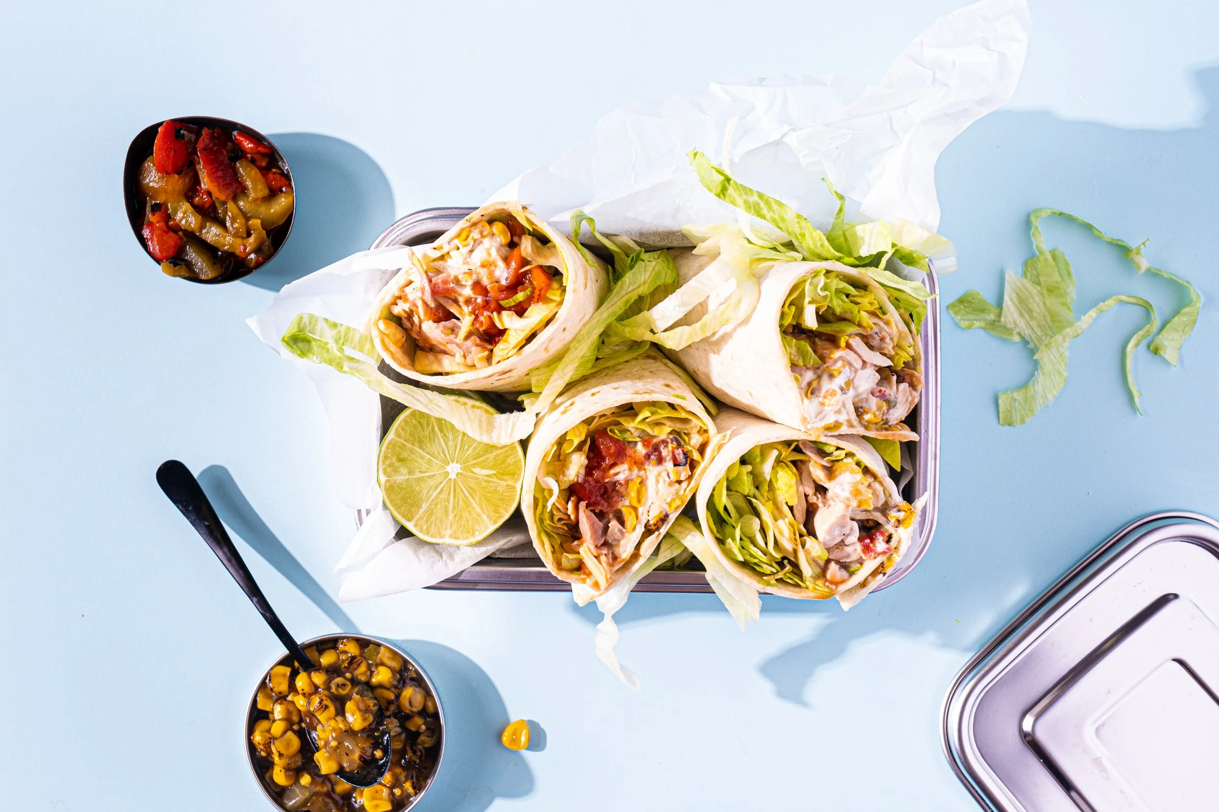 Bilde av fire wrap-sandwicher fylt med grønnsaker, kylling og ost, servert med sitronskiver og coleslaw, samt to små boller med mais og svartbønner