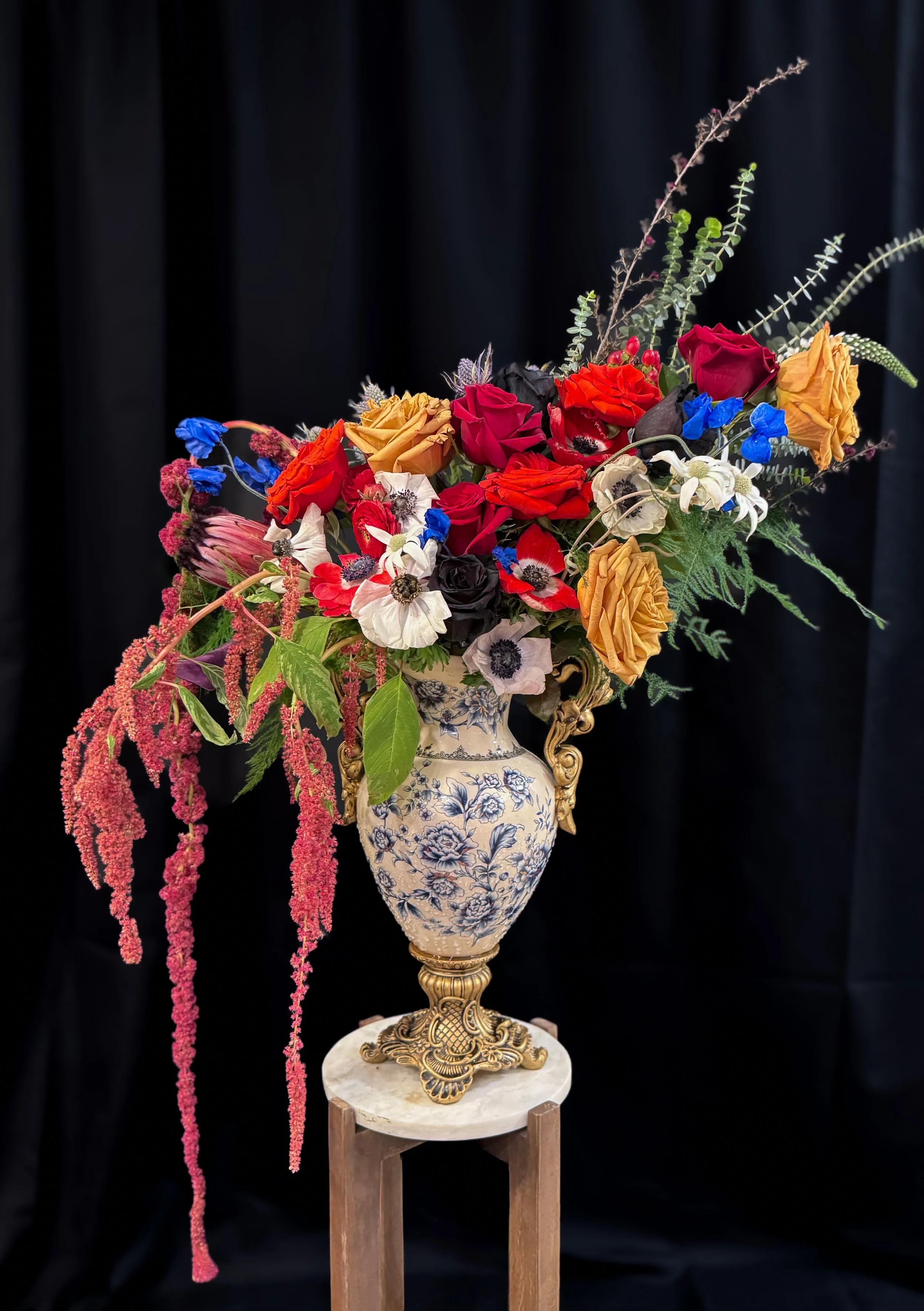 A large floral arrangement with red, yellow, white, and blue flowers in a decorative blue and white vase, placed on a small wooden stand against a dark background.