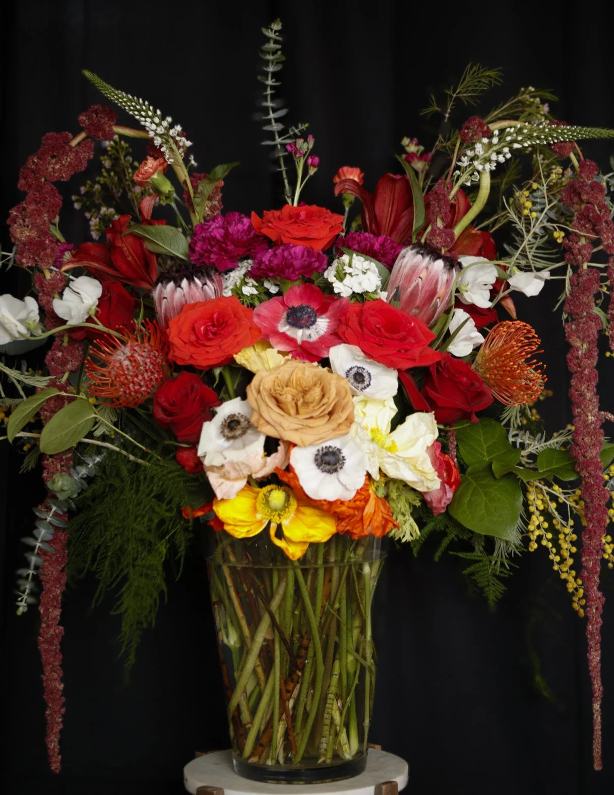 A colorful bouquet of various flowers in a glass vase, featuring red, orange, yellow, white, and purple flowers, with green foliage, set against a black background.