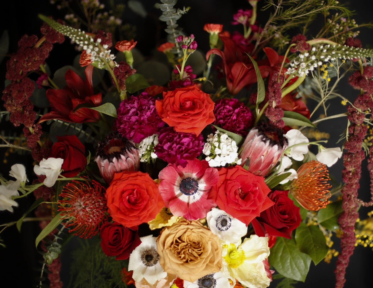 A colorful bouquet of various flowers including roses, anemones, proteas, and other blossoms, arranged with green foliage.