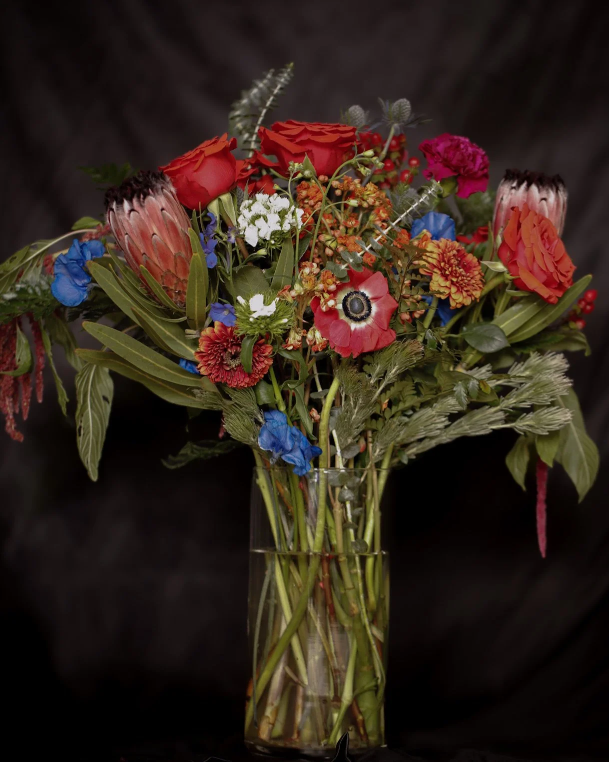 Colorful mixed flower bouquet in a glass vase against a dark background.