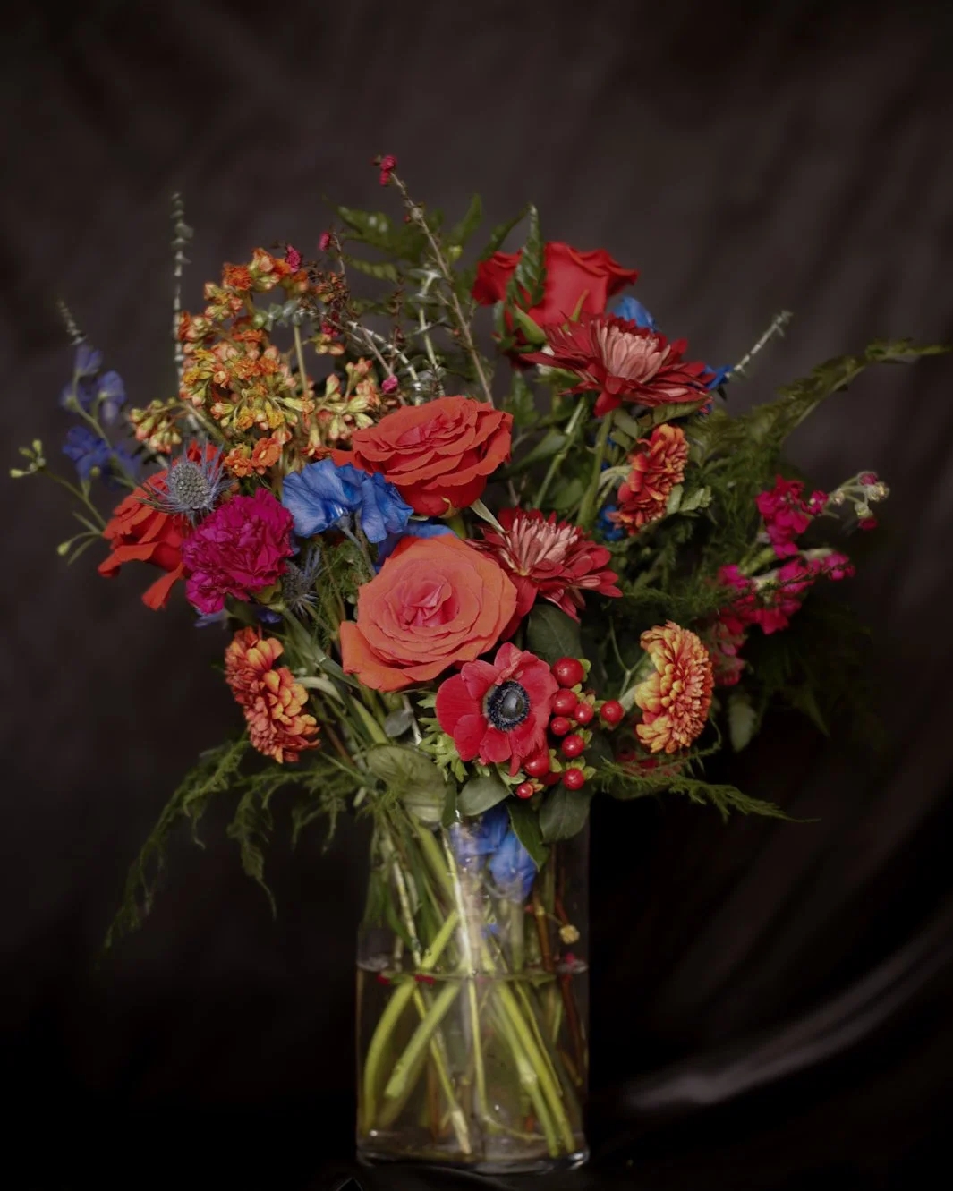 A colorful bouquet of mixed flowers including roses, daisies, and other blooms in a glass vase against a dark background.
