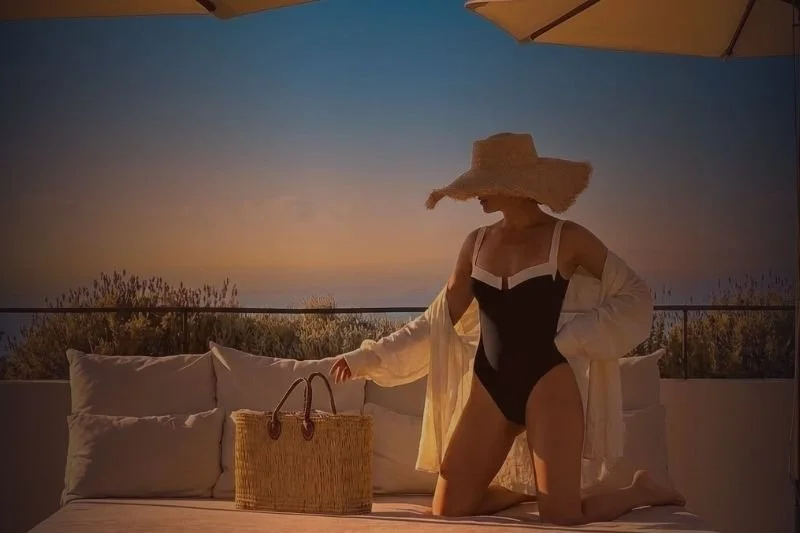 Woman in a black swimsuit and large sunhat on a balcony at sunset, with pillows and a straw bag, under a yellow umbrella.