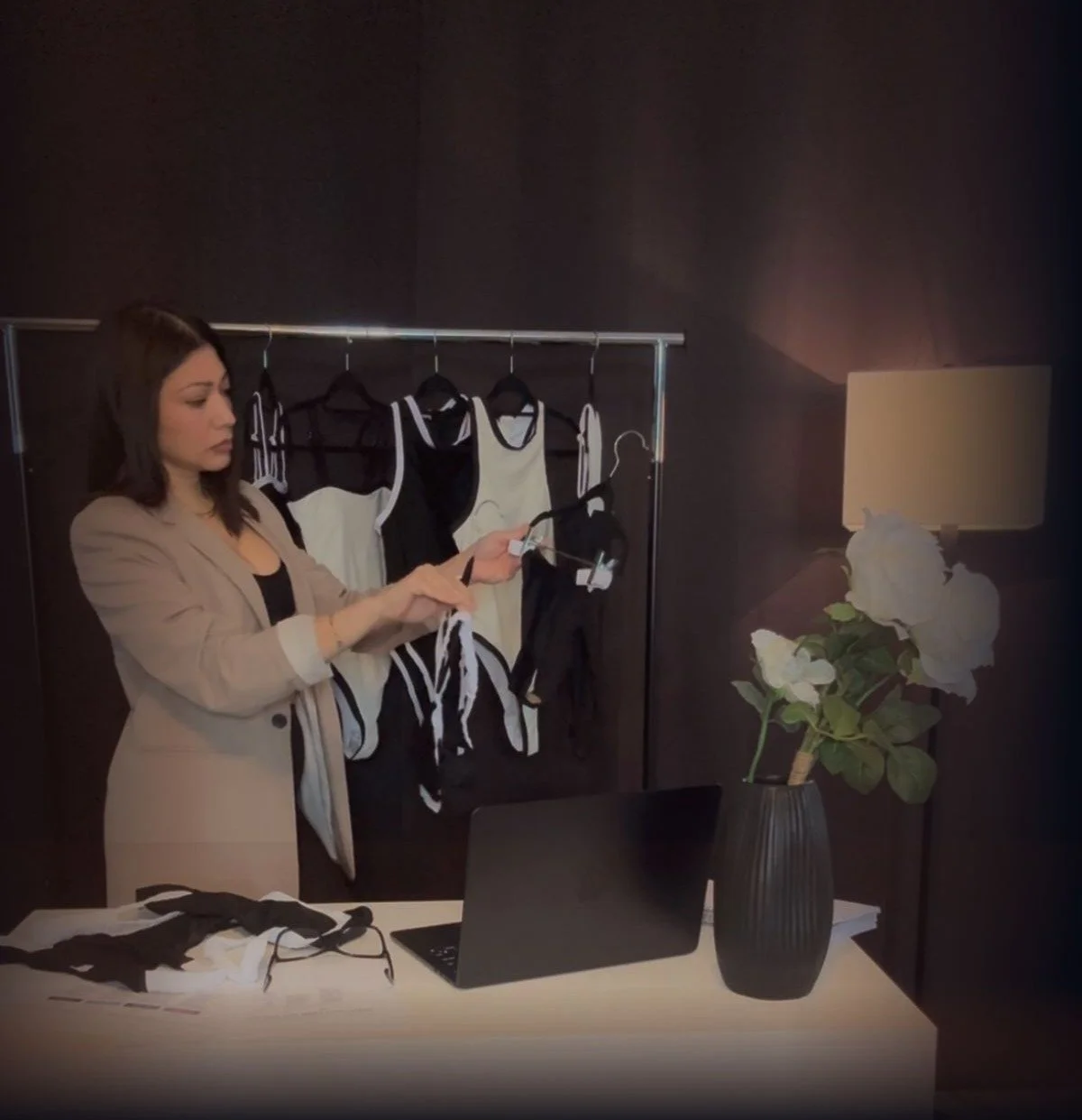 A woman in a beige blazer looks at a clothing tag on a black strap in a room with a clothing rack of black and white garments, a brown vase with white flowers, and a table with a black laptop and glasses.